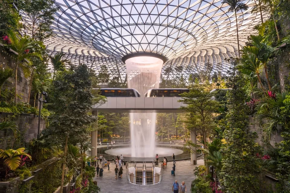 The Jewel at Changi Airport, showcasing the stunning indoor waterfall surrounded by lush greenery and a modern glass dome, with a sky bridge and people walking below.