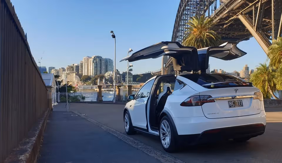 A white Tesla Model X with its falcon wing doors open, parked near the iconic Harbour Bridge.