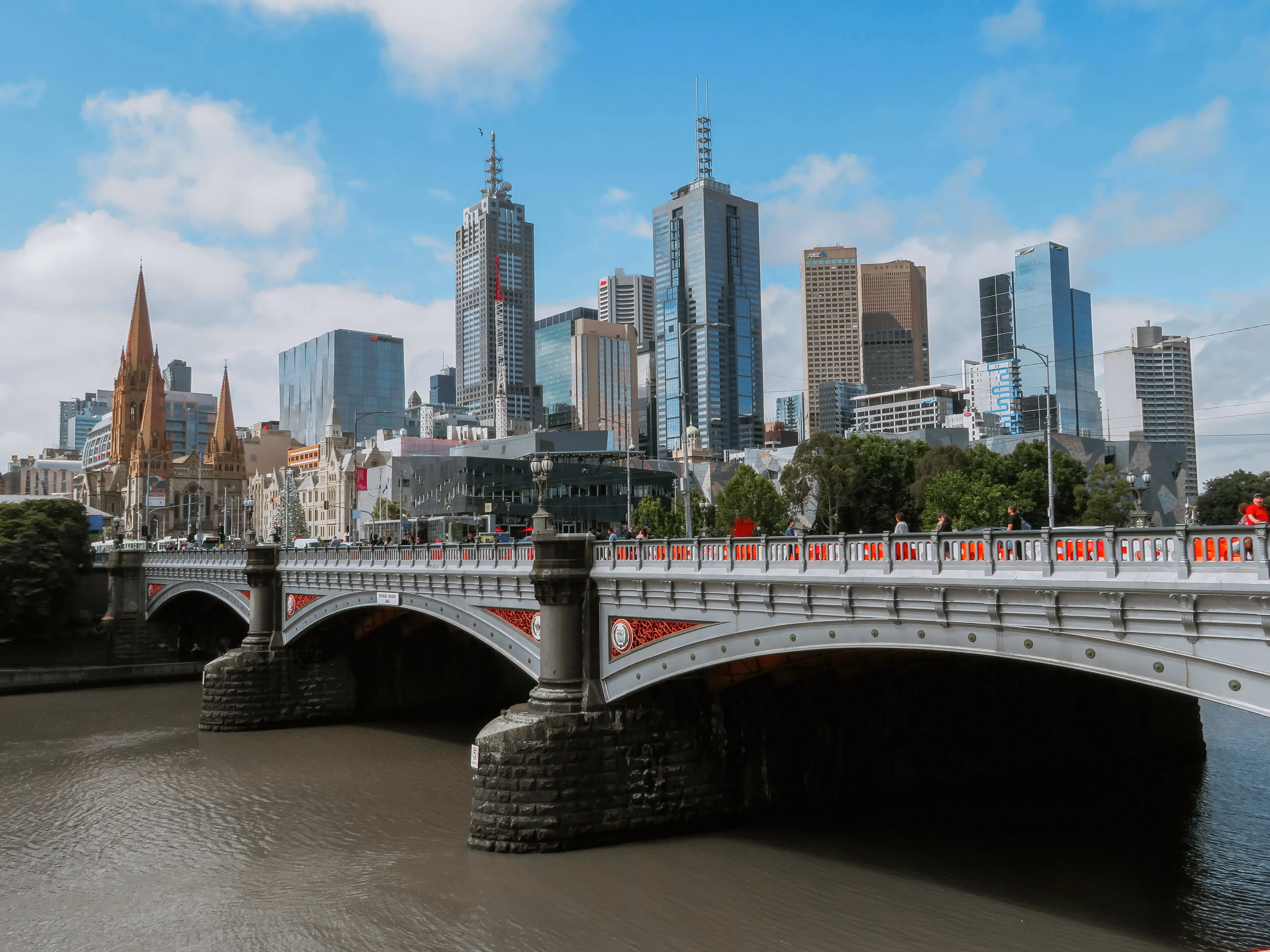 A view of Melbourne's skyline with a mix of modern skyscrapers and historic buildings, featuring St Paul's Cathedral's spires and the ornate Princes Bridge spanning the Yarra River under a clear blue sky.