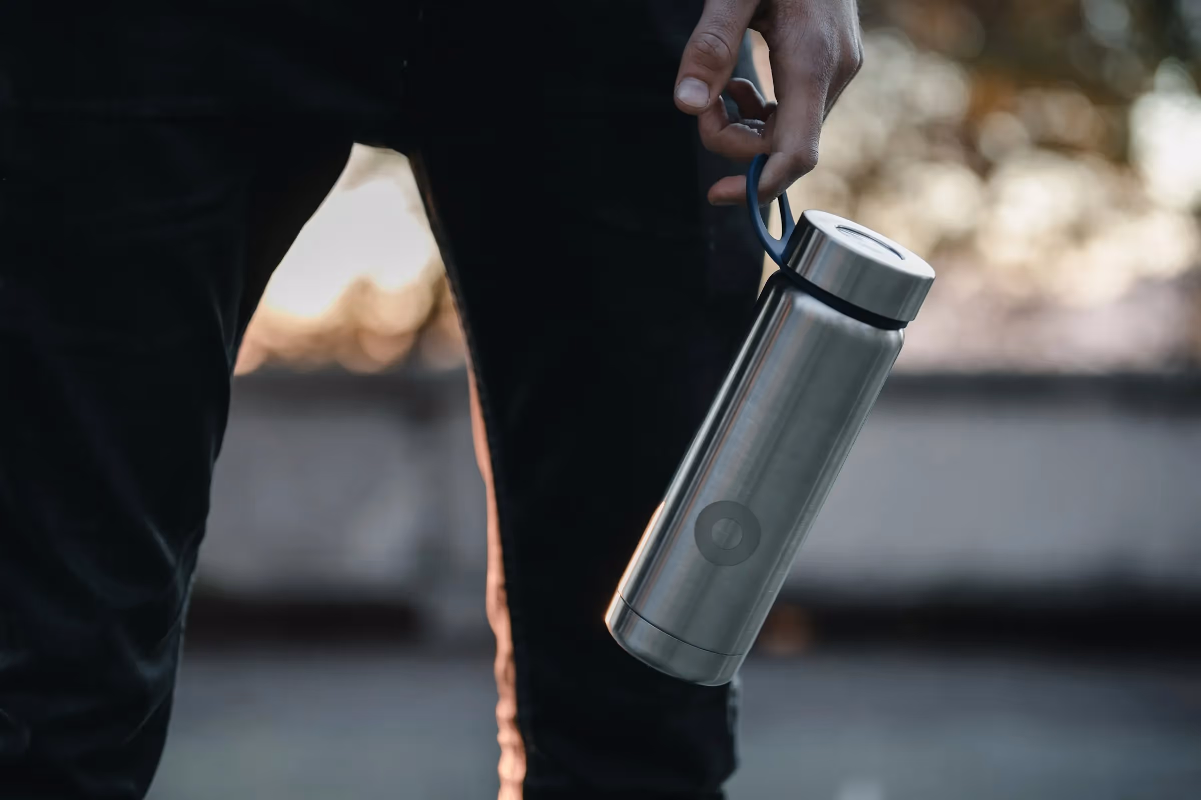 A person holding a stainless steel reusable water bottle by its loop, with the background softly blurred and natural sunlight creating a warm atmosphere.