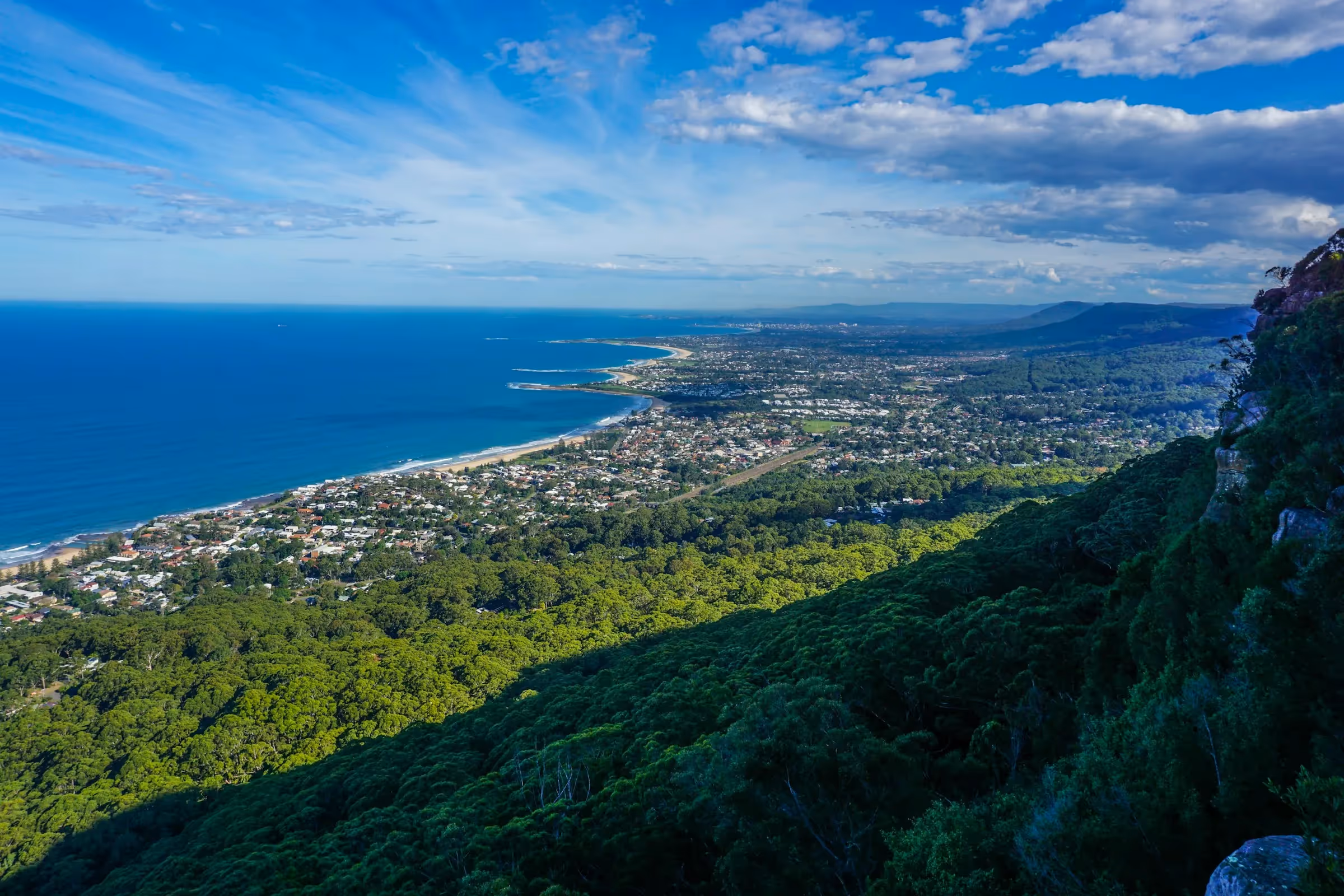 A breathtaking aerial view of Wollongong, showcasing its stunning coastline, lush green landscape, and the sprawling cityscape under a clear blue sky.