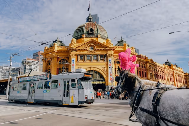 A tram passes by the iconic Flinders Street Station in Melbourne, with a horse-drawn carriage in the foreground.