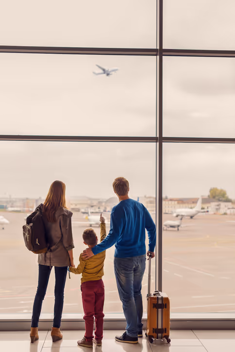 A family of three, including a mother, father, and young child, stands by a large airport window, watching an airplane take off.