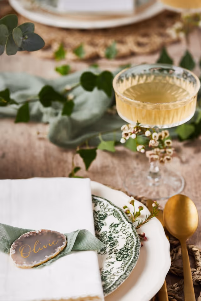 A beautifully set wedding table with a green and white plate, linen napkin with a name tag, gold spoon, and a glass of champagne, accented with greenery.