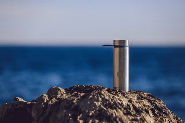 A stainless steel reusable water bottle sitting on a rocky surface with the ocean in the background.