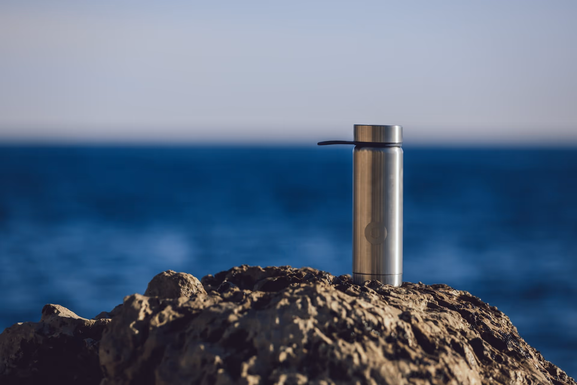 A stainless steel reusable water bottle sitting on a rocky surface with the ocean in the background.