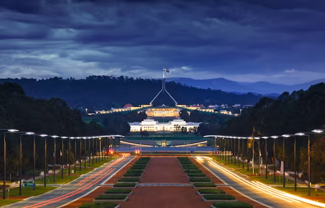 A night-time view of Canberra's Parliament House, illuminated against a backdrop of dark clouds and the surrounding landscape.