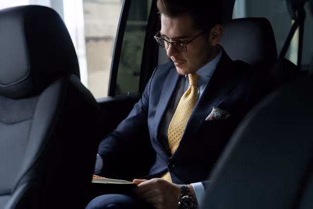 A corporate professional, seated in the back of a luxury vehicle, working while being driven by a chauffeur.