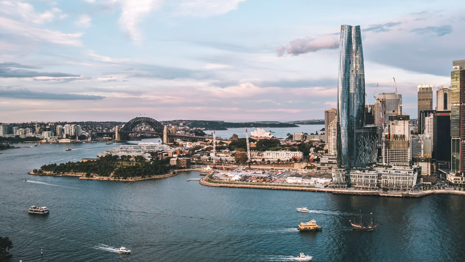 A view of Sydney Harbour, featuring the Opera House, ferries, and boats moving across the water, with the city skyline in the background on a sunny day.