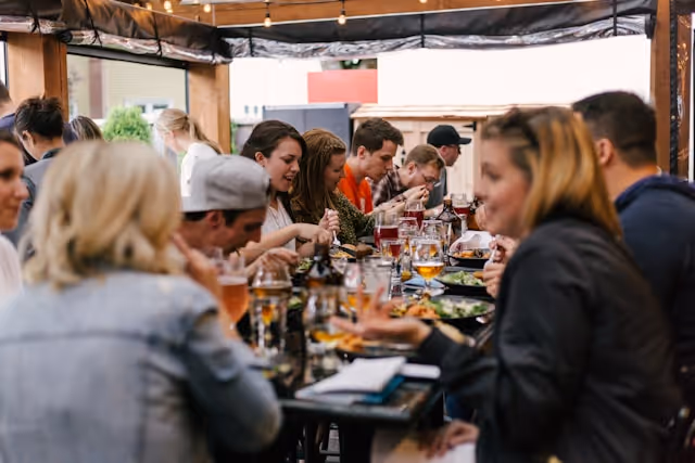 Group of colleagues enjoying a meal outdoors, gathered around a long table with food, drinks, and conversation under string lights.