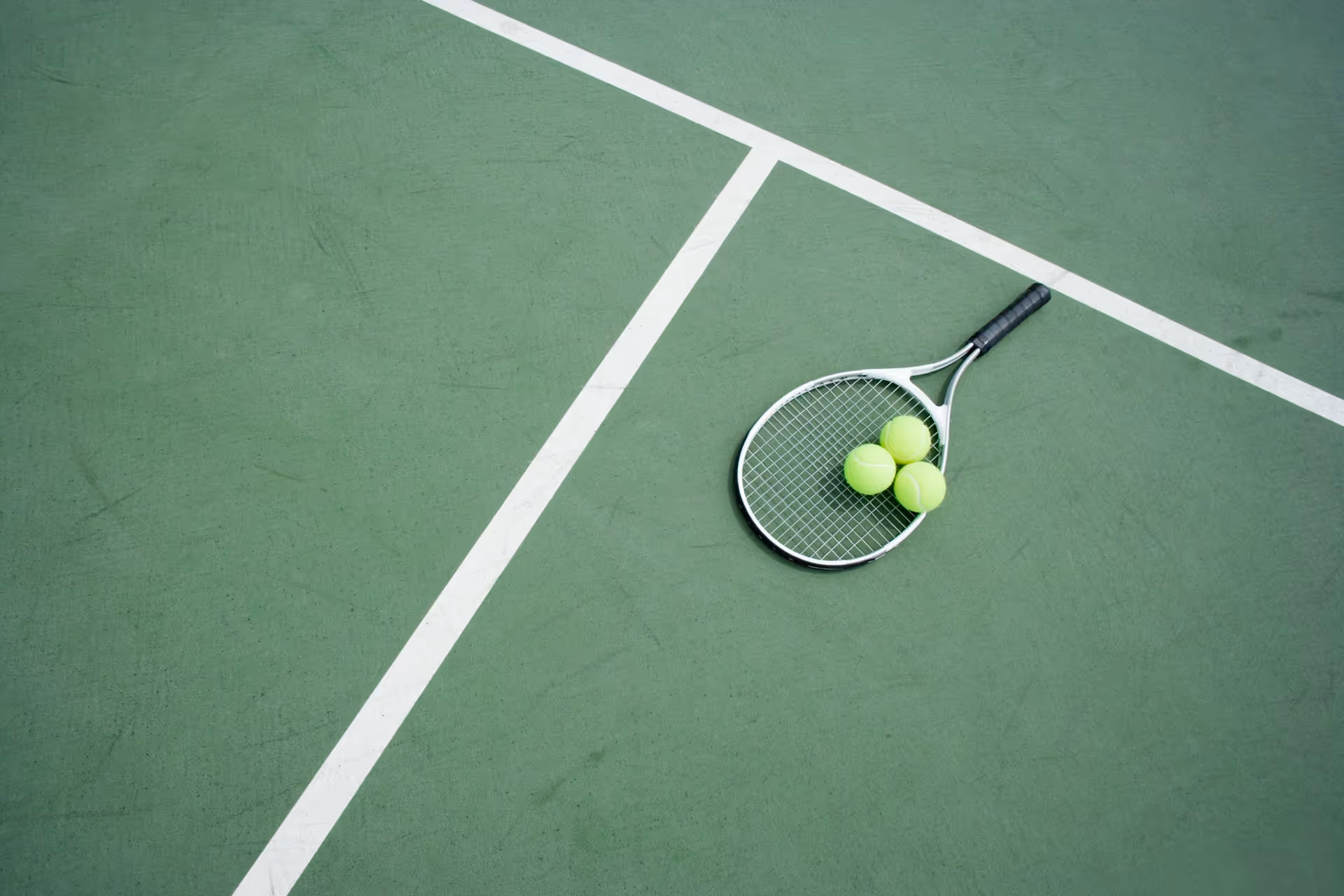 A tennis racket and three tennis balls lying on a green tennis court near the white boundary lines.