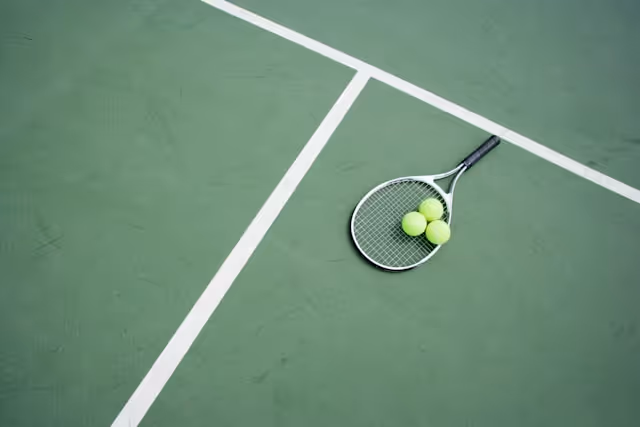 A tennis racket and three tennis balls lying on a green tennis court near the white boundary lines.