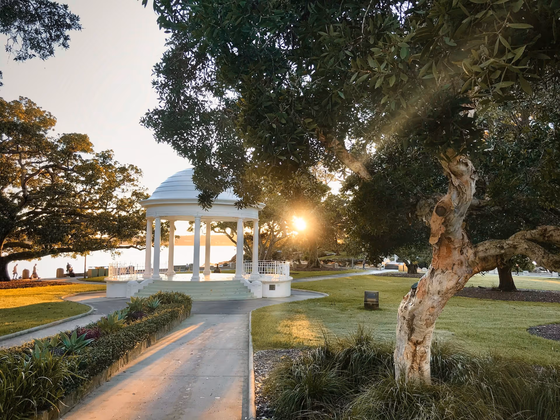 Sunlight shining through trees at Balmoral Beach, with the white rotunda in the foreground and people walking along the waterfront path.