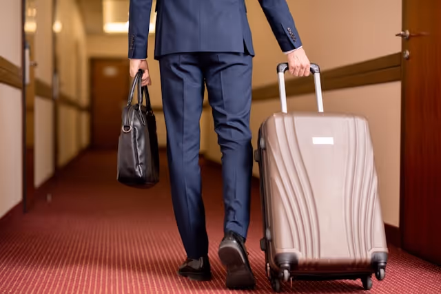 Businessman in a navy suit walking down a hotel corridor, carrying a black leather bag in one hand and pulling a brown suitcase with the other.