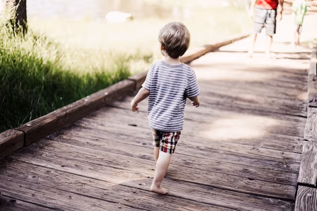 Young child walking barefoot on a wooden pathway surrounded by grass.
