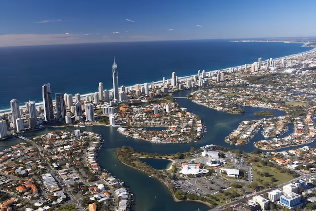 Aerial view of the Gold Coast, featuring high-rise buildings along the coastline, waterways winding through residential areas, and the expansive blue ocean under a clear sky.