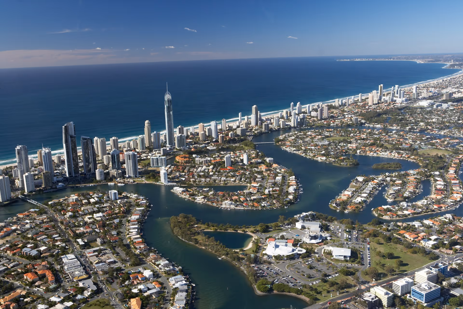 Aerial view of the Gold Coast, featuring high-rise buildings along the coastline, waterways winding through residential areas, and the expansive blue ocean under a clear sky.