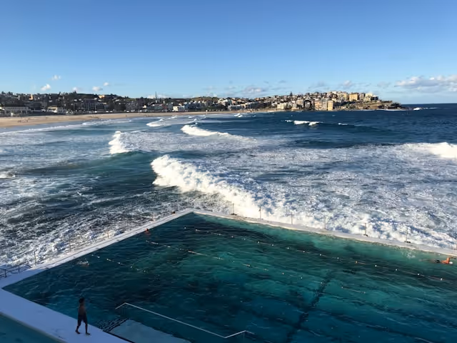 Bondi Icebergs Pool overlooking the ocean, with waves crashing against the edge of the pool and Bondi Beach in the background under a bright blue sky.