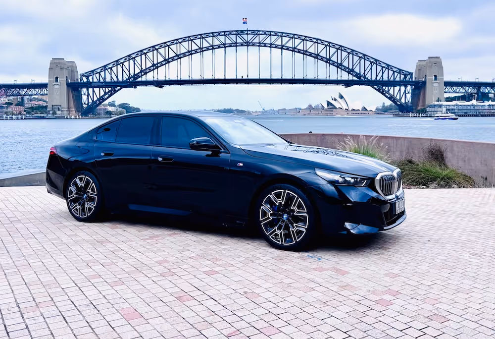 BMW i5 parked on a paved area with the Sydney Harbour Bridge and Opera House in the background.