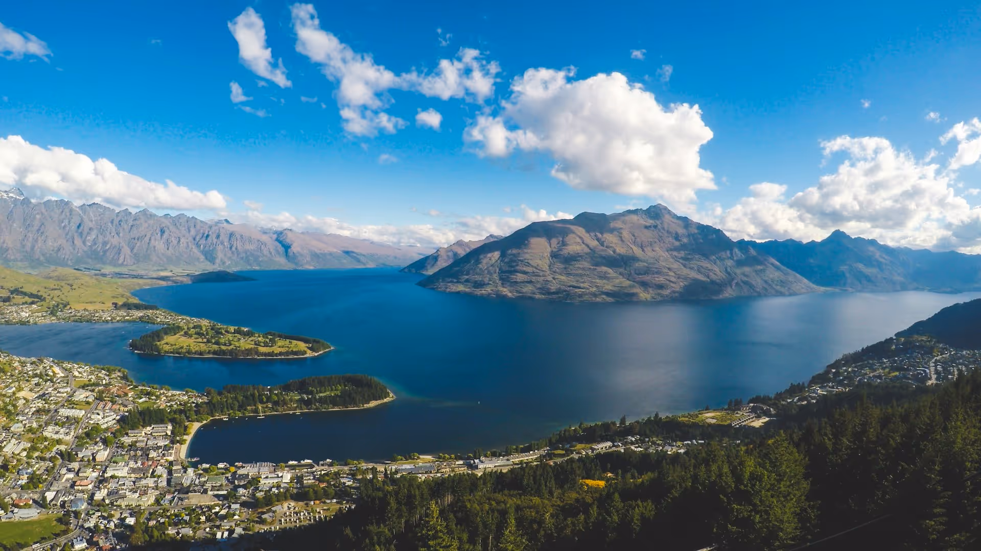 A panoramic view of Queenstown, New Zealand, showcasing Lake Wakatipu, under a bright blue sky with scattered clouds.