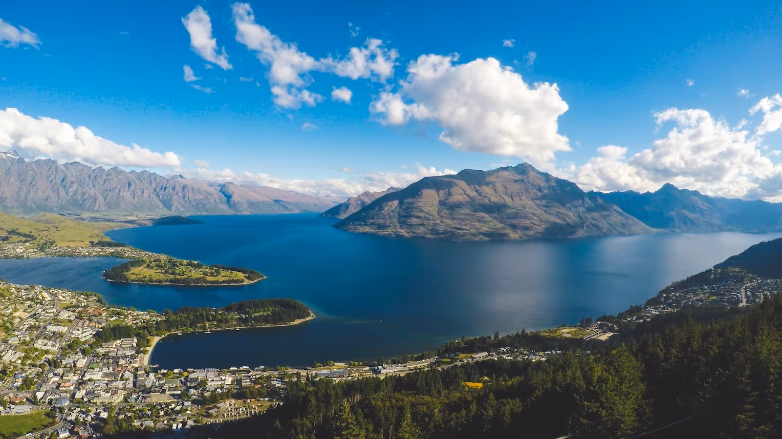 A panoramic view of Queenstown, New Zealand, showcasing Lake Wakatipu, under a bright blue sky with scattered clouds.