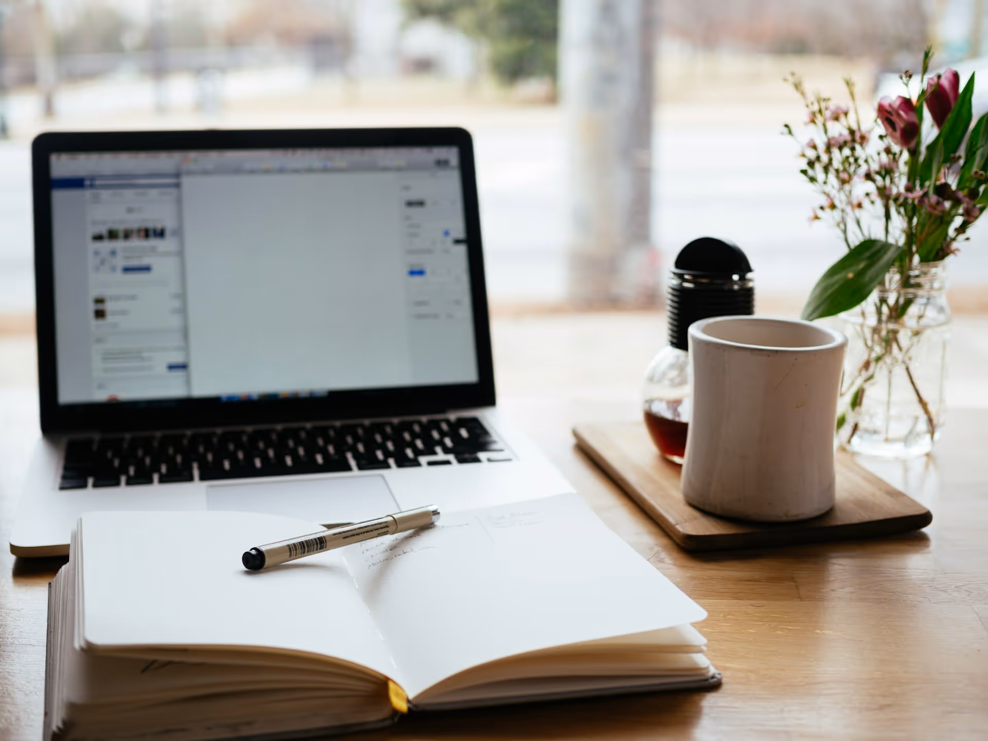 An open notebook with a pen rests on a desk next to a laptop, a coffee mug, and a small vase of flowers, creating a workspace setting by a window.