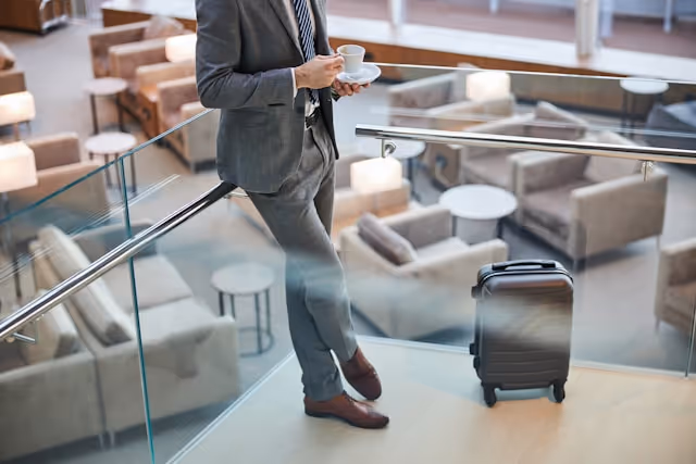 Business traveller in a grey suit, holding a cup of coffee with a carry-on suitcase.