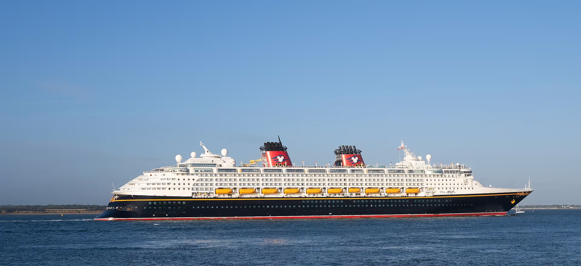 A Disney cruise ship sailing across calm waters under a clear blue sky, featuring the iconic red and black funnels with Mickey Mouse logos.