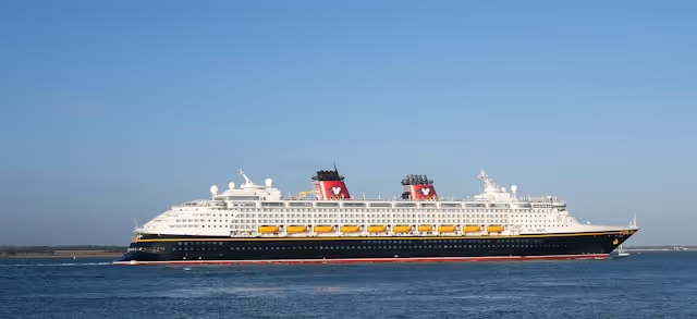 A Disney cruise ship sailing across calm waters under a clear blue sky, featuring the iconic red and black funnels with Mickey Mouse logos.
