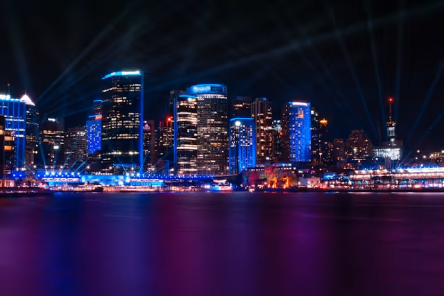 A stunning view of Sydney’s skyline at night during Vivid Sydney, with illuminated skyscrapers, vibrant blue and purple lights, and beams of light radiating into the sky.
