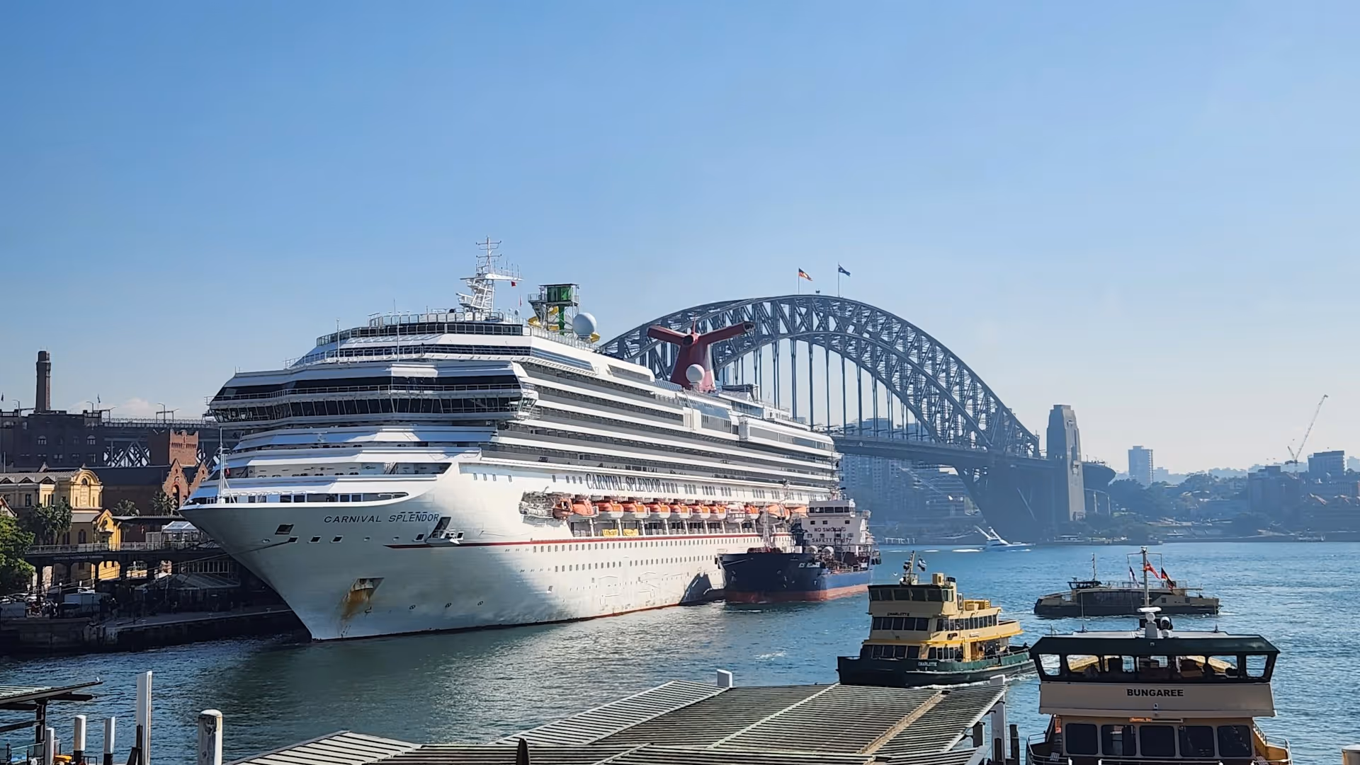 A large cruise ship, the Carnival Splendor, docked at Sydney's Overseas Passenger Terminal with the Harbour Bridge in the background, alongside ferries.