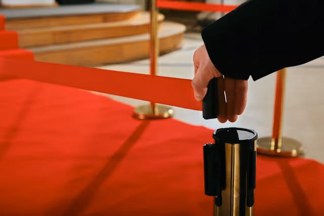 A hand in a black suit opens a red barrier on a red carpet, leading to an event.