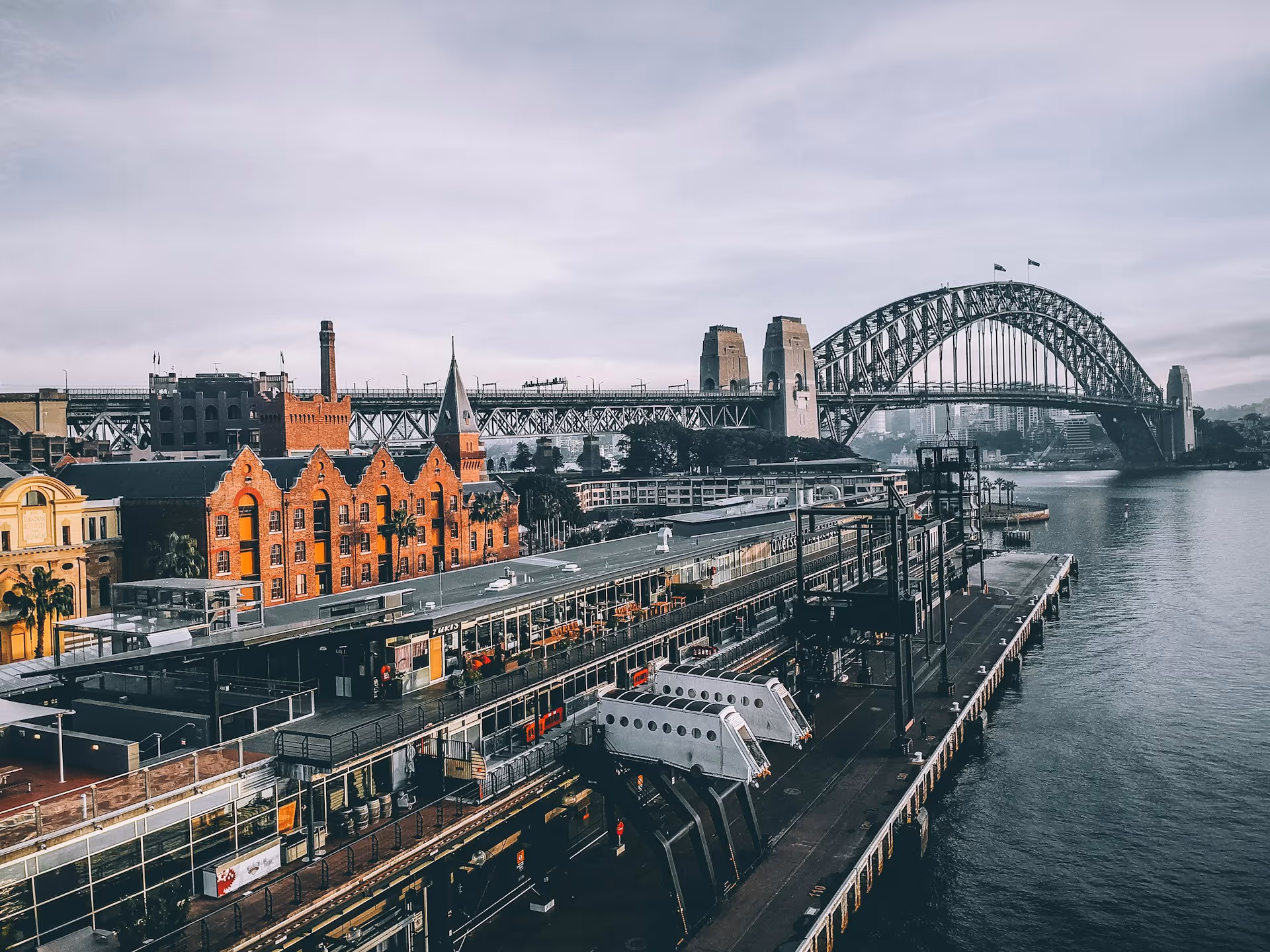 View of Sydney's Overseas Passenger Terminal with historic buildings and the Sydney Harbour Bridge in the background under a cloudy sky.