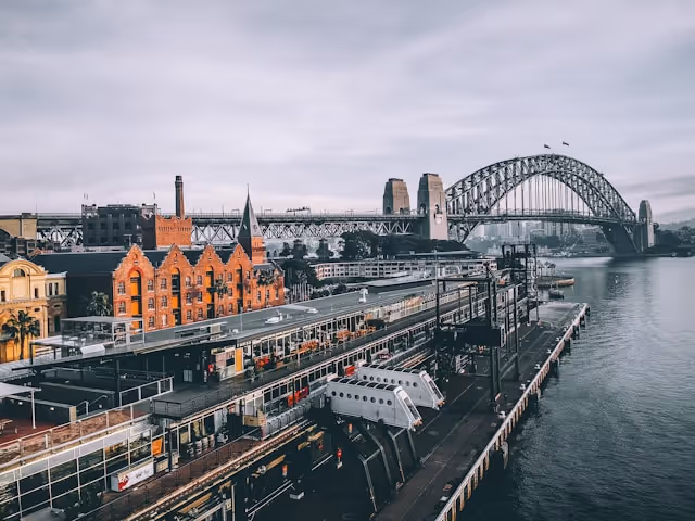 View of Sydney's Overseas Passenger Terminal with historic buildings and the Sydney Harbour Bridge in the background under a cloudy sky.