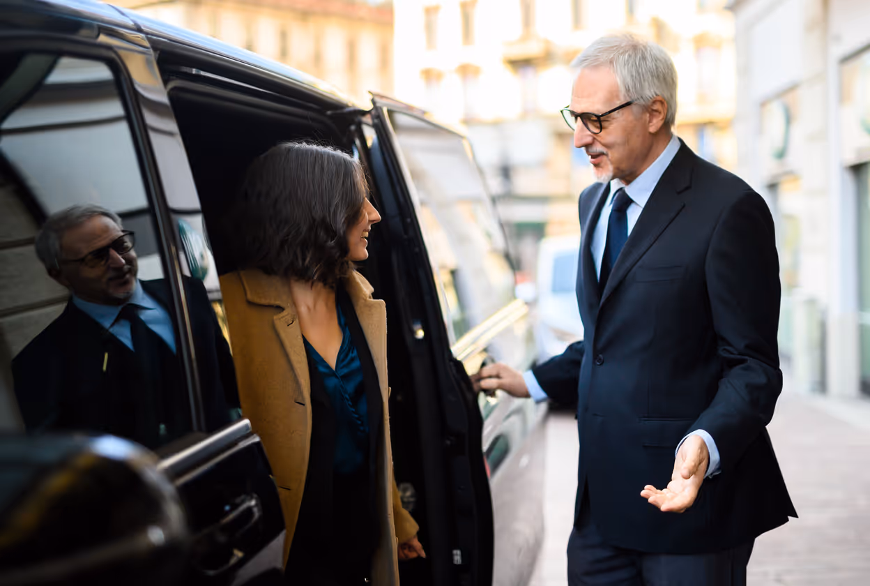 A professional chauffeur greets a female passenger while opening the door of a premium black van.