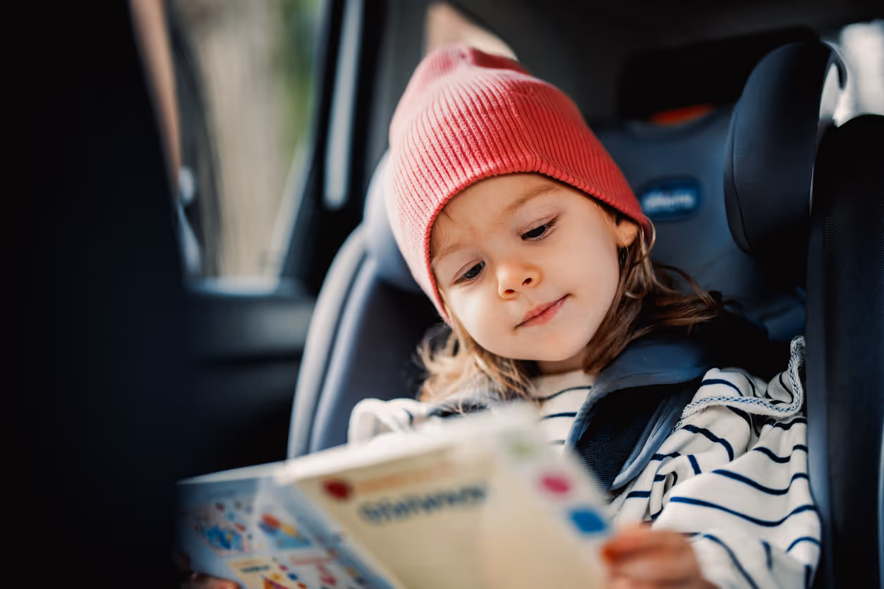Young child seated safely in a car seat, wearing a red beanie and reading a colourful book during a car journey.