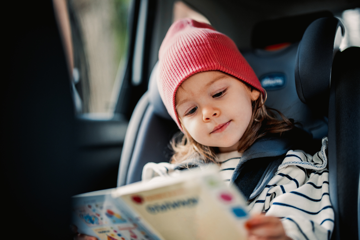 Young child seated safely in a car seat, wearing a red beanie and reading a colourful book during a car journey.