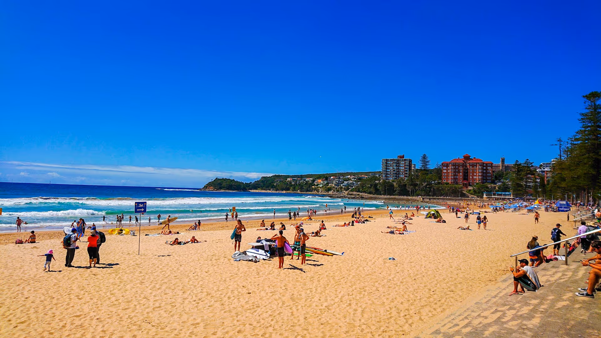 People relaxing and walking along the sand at Manly Beach on a bright sunny day, with clear blue skies and buildings in the background.