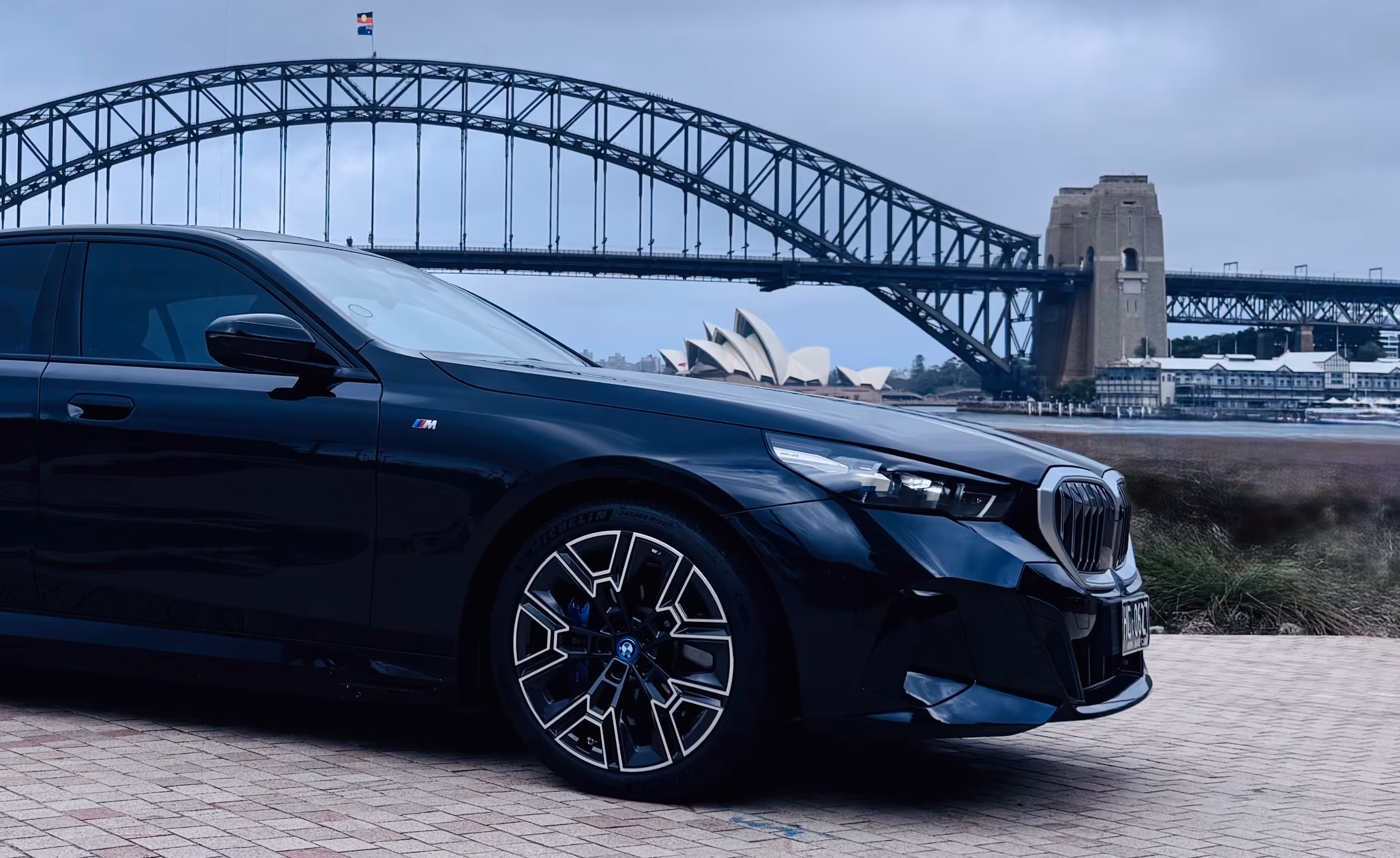 BMW i5 parked near Sydney Harbour with the Sydney Harbour Bridge and Opera House in the background on a cloudy day.