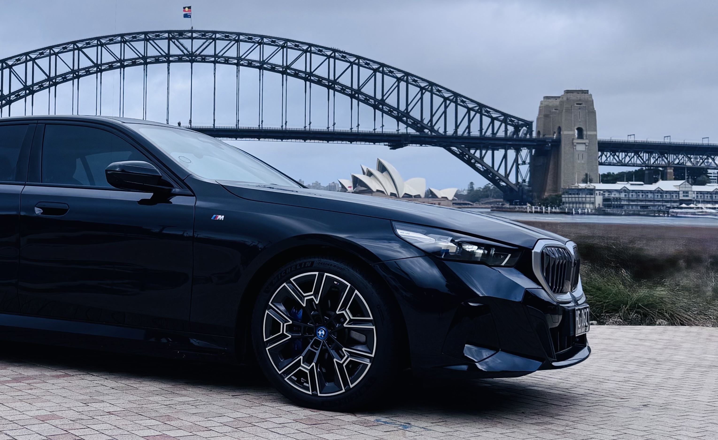 BMW i5 parked near Sydney Harbour with the Sydney Harbour Bridge and Opera House in the background on a cloudy day.
