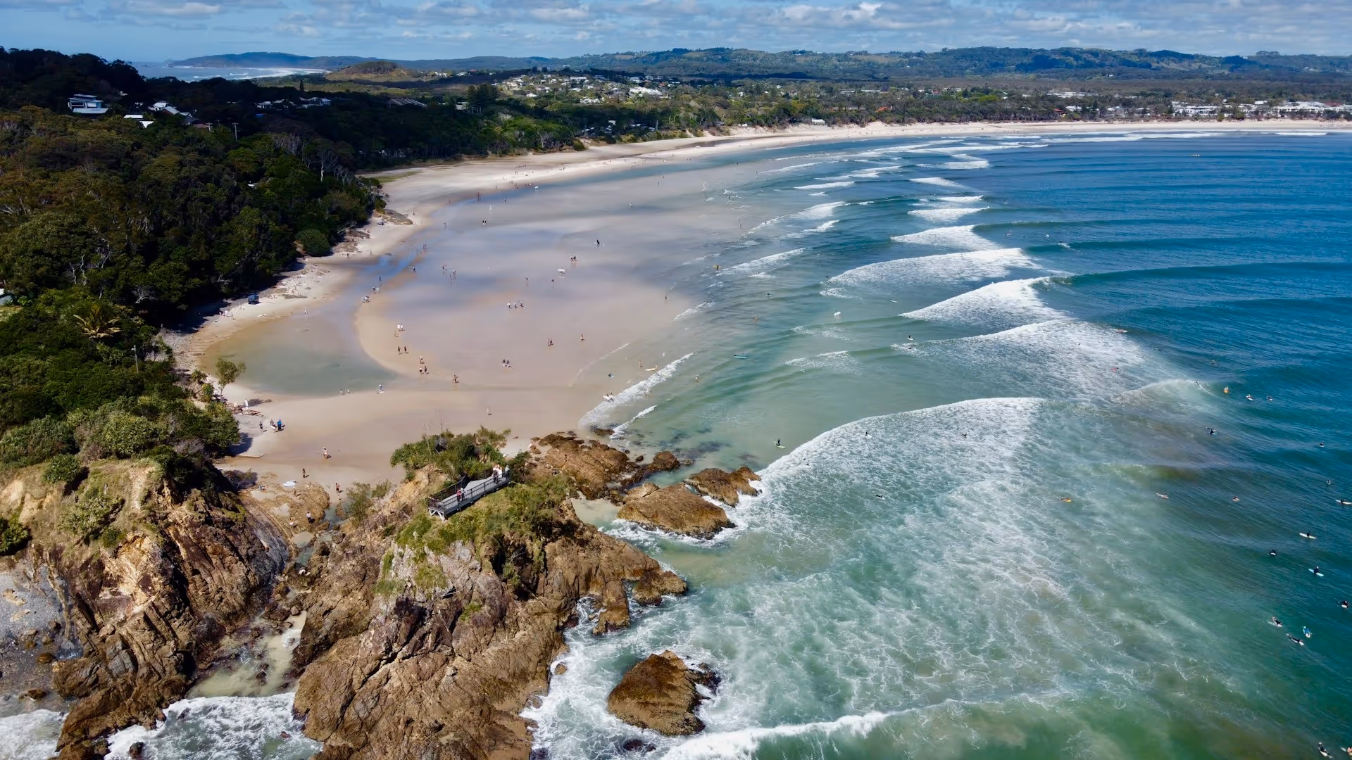 Aerial view of The Pass, Byron Bay, showcasing a sandy beach, clear blue ocean with surfers, rocky headland, and surrounding lush greenery