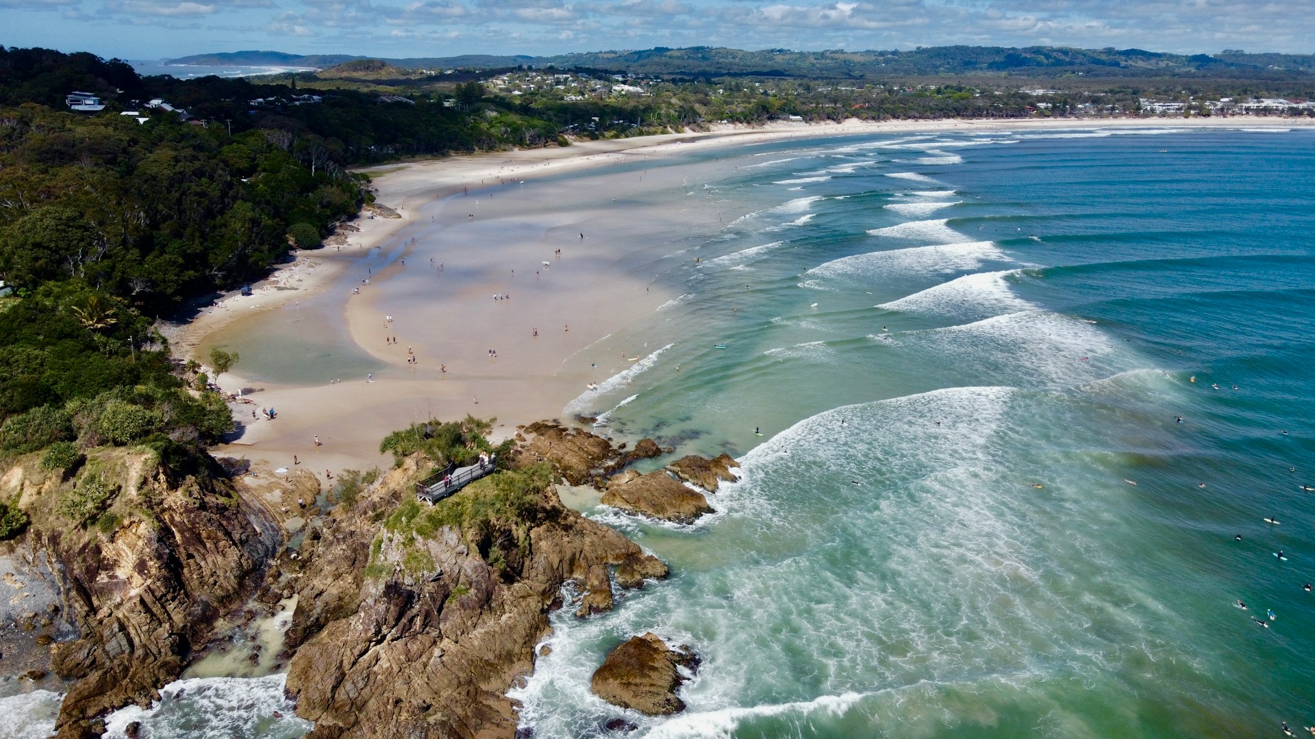 Aerial view of The Pass, Byron Bay, showcasing a sandy beach, clear blue ocean with surfers, rocky headland, and surrounding lush greenery