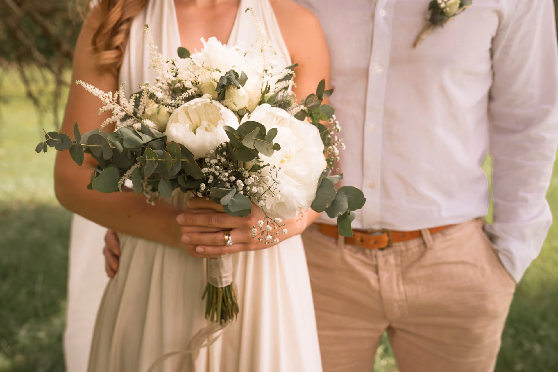A bride and groom stand together outdoors, with the bride holding a bouquet of white peonies and greenery.