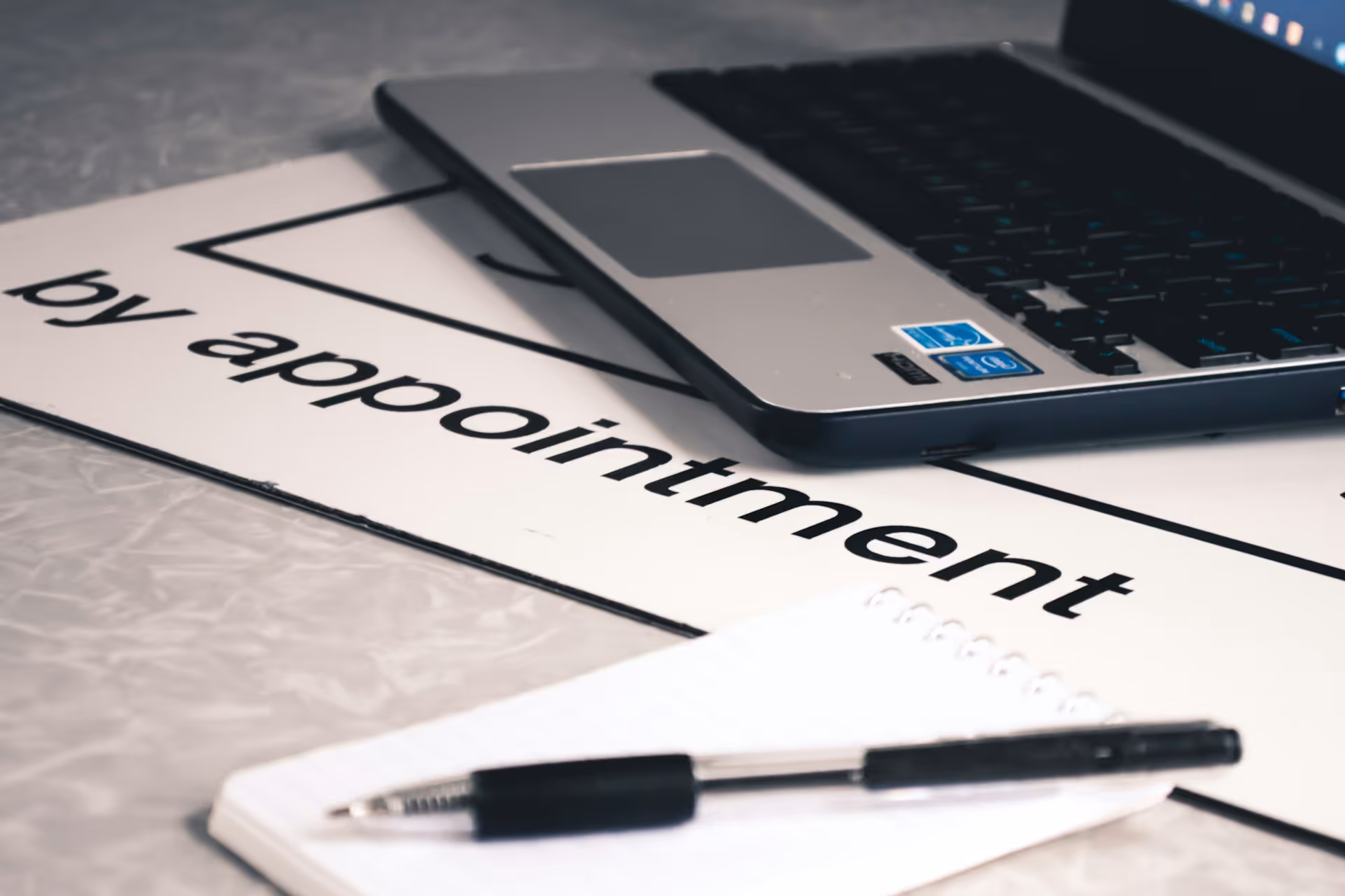 Close-up of a sign reading "by appointment" alongside a laptop and notepad, representing scheduled commitments and the convenience of having a chauffeur wait during appointments or events.