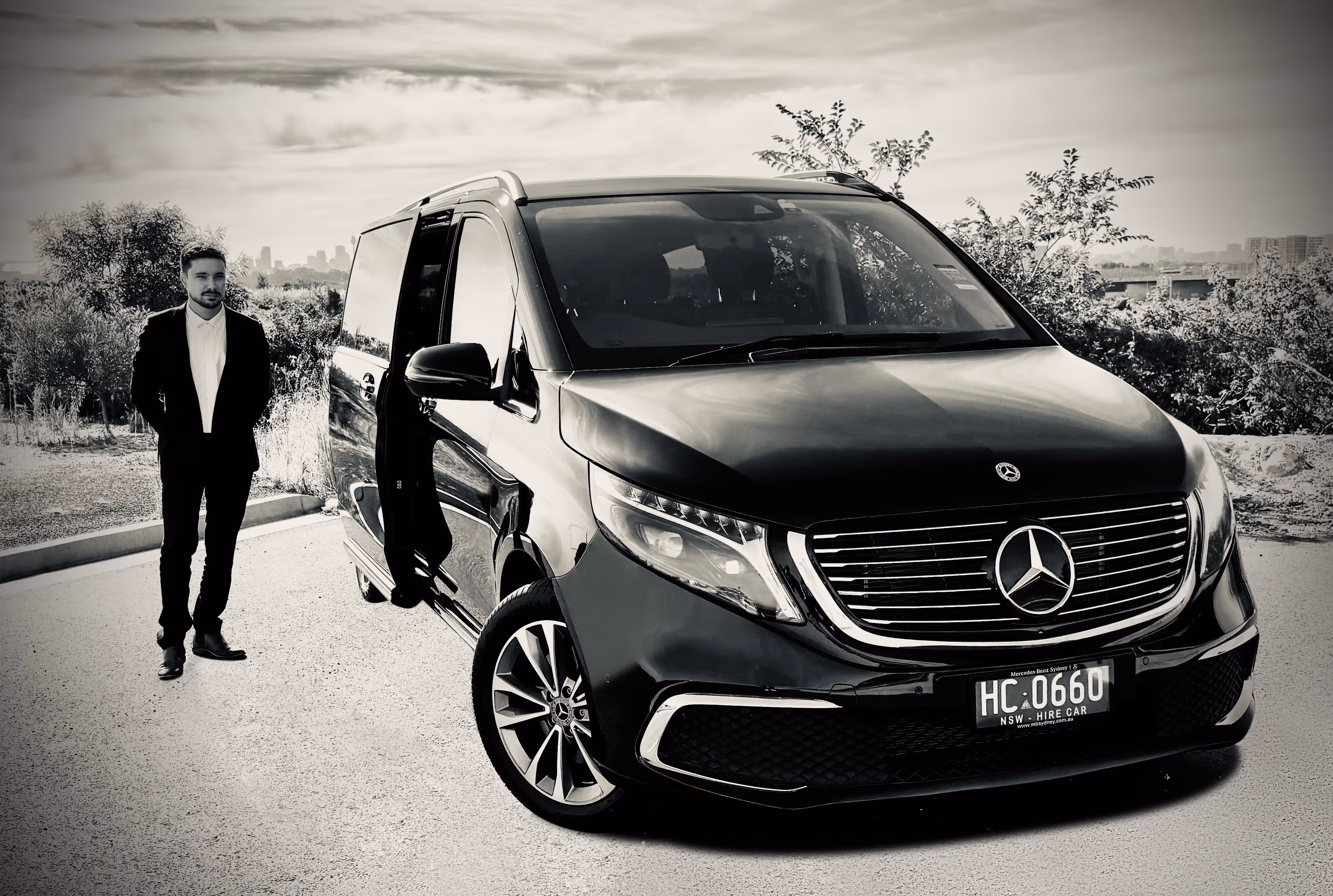 An Evoke chauffeur stands beside a black Mercedes-Benz EQV with its sliding door open, parked in an outdoor setting with city buildings visible in the background.