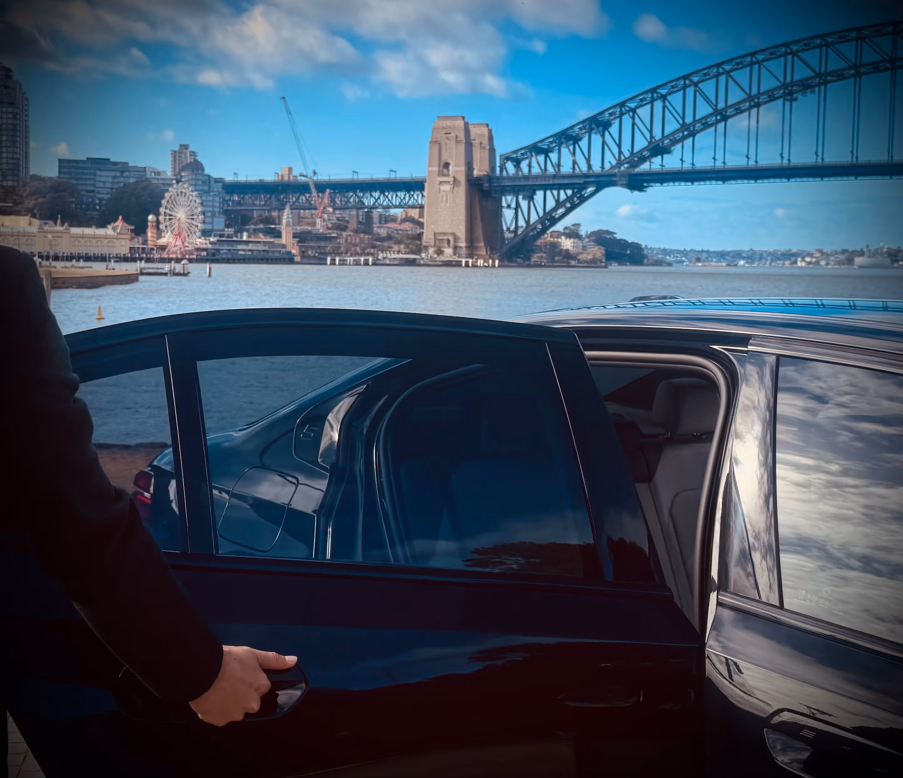 Chauffeur opening the rear door of a BMW with Sydney Harbour Bridge and Luna Park visible in the background.