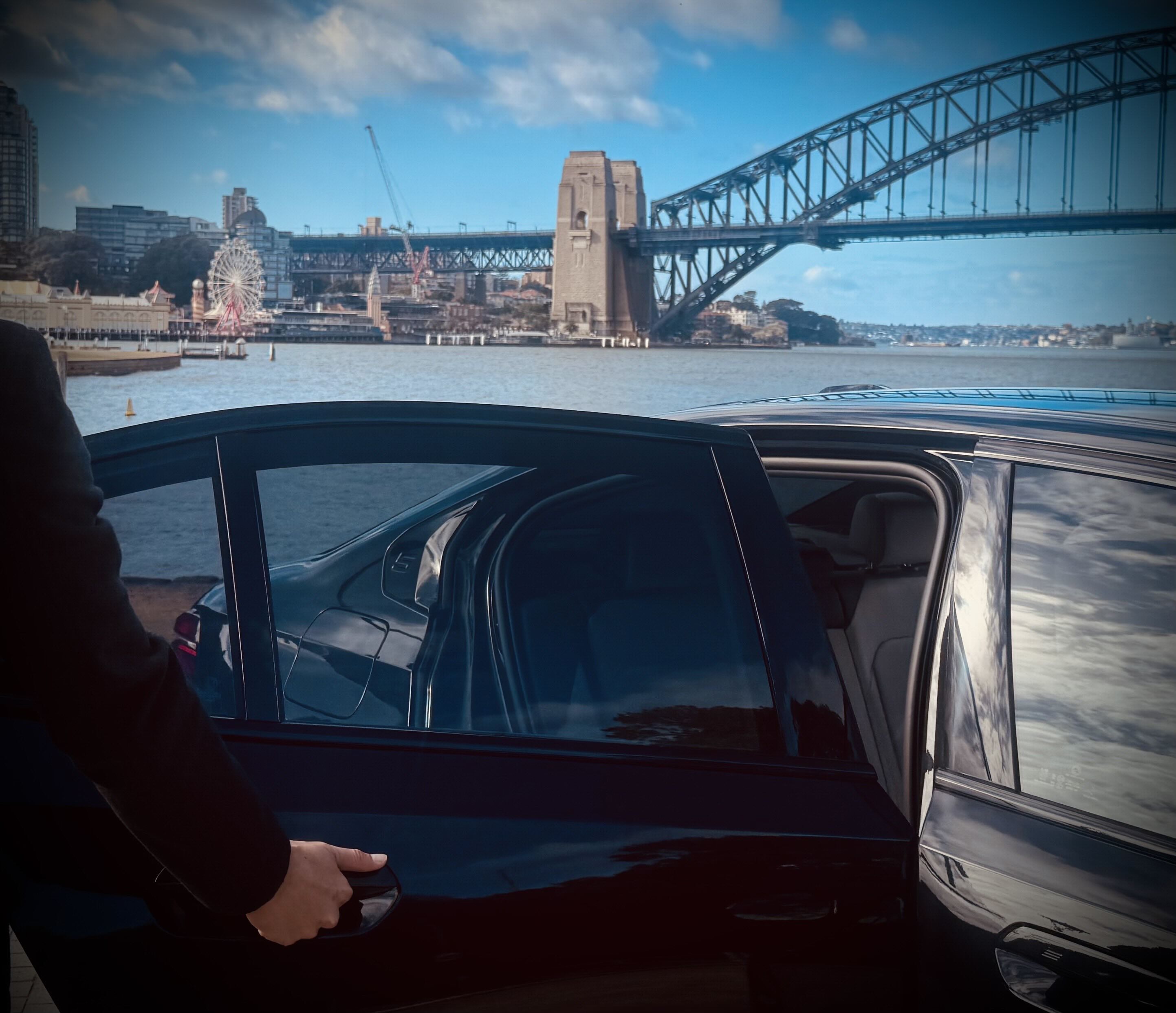 Chauffeur opening the rear door of a BMW with Sydney Harbour Bridge and Luna Park visible in the background.
