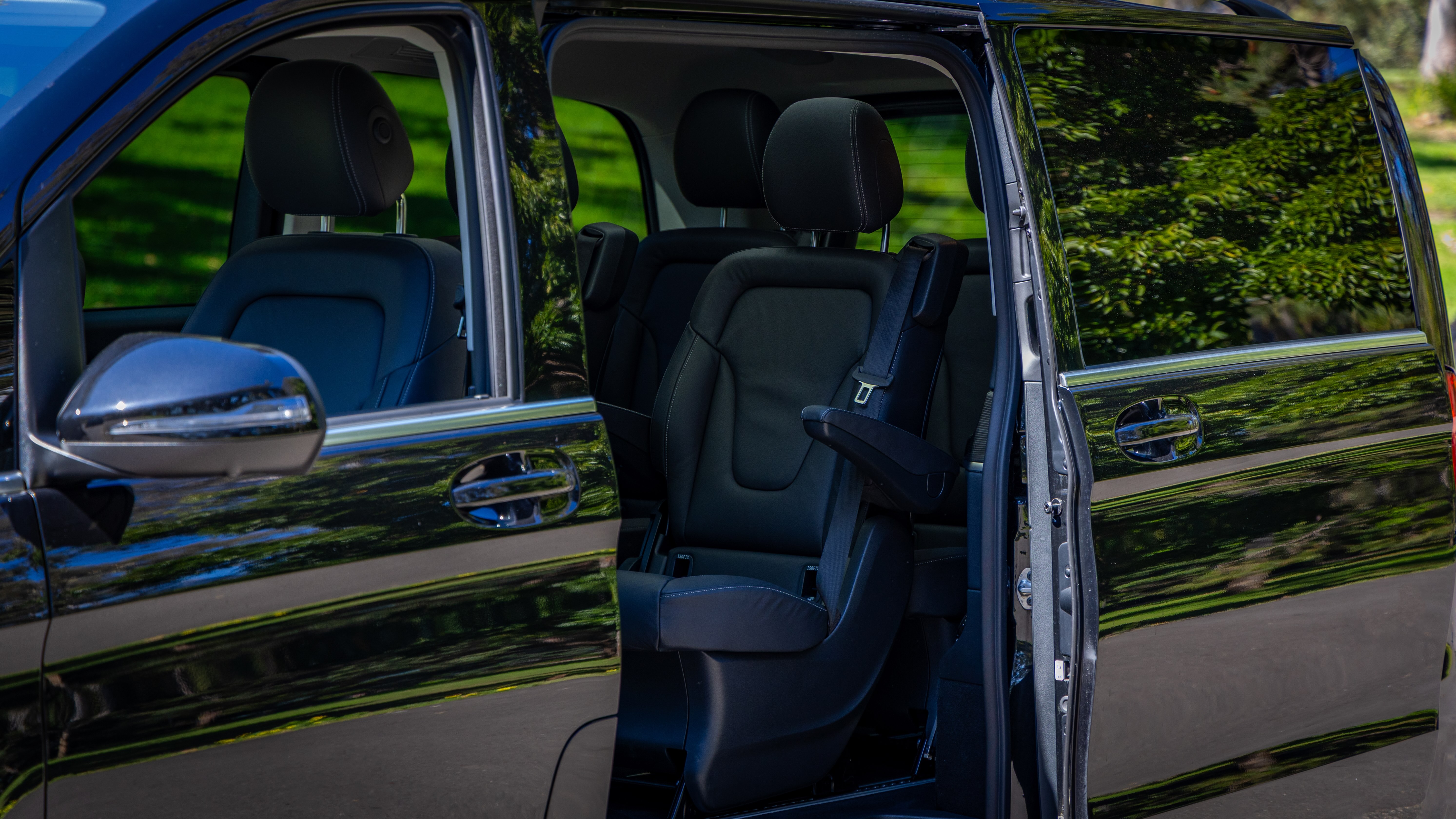 Mercedes EQV interior featuring black leather captain seats with armrests, captured through the open sliding door with reflections of trees on the exterior.