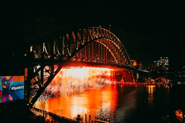 Fireworks light up the Sydney Harbour Bridge during New Year’s Eve celebrations, with reflections on the water and crowds gathered below.