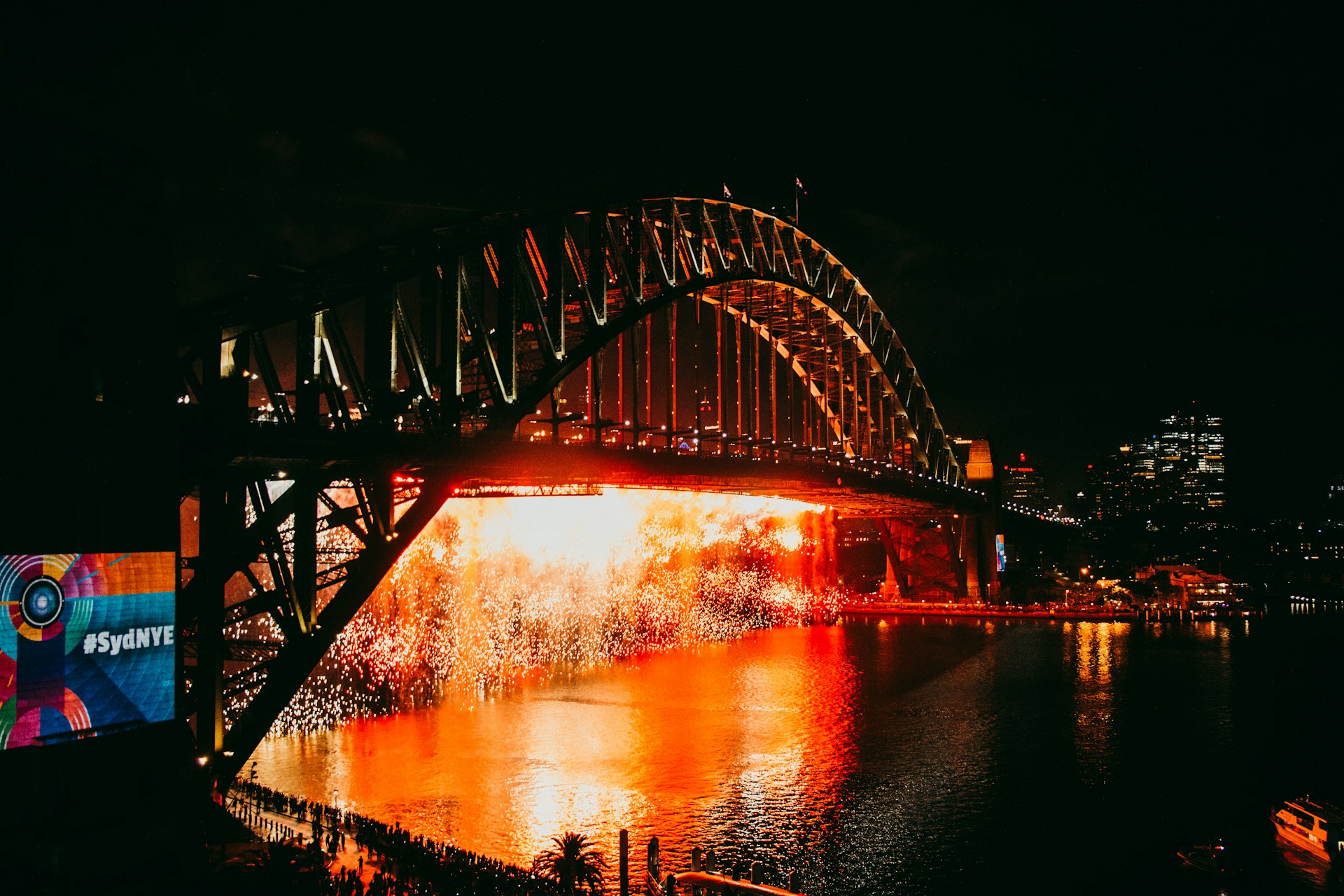Fireworks light up the Sydney Harbour Bridge during New Year’s Eve celebrations, with reflections on the water and crowds gathered below.
